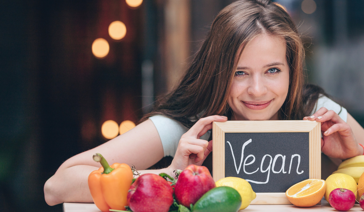 a girl holding a mini black board with vegan written on it and vegetables put on a counter in front of her