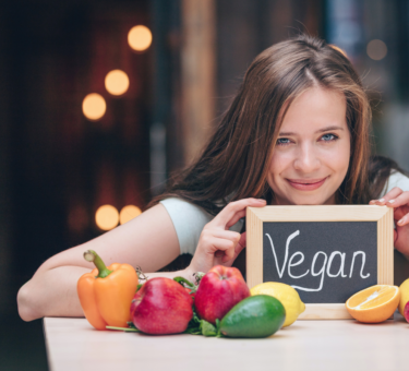 a girl holding a mini black board with vegan written on it and vegetables put on a counter in front of her