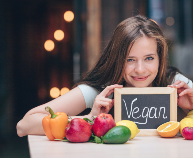 a girl holding a mini black board with vegan written on it and vegetables put on a counter in front of her