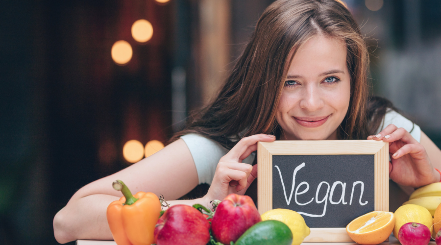 a girl holding a mini black board with vegan written on it and vegetables put on a counter in front of her